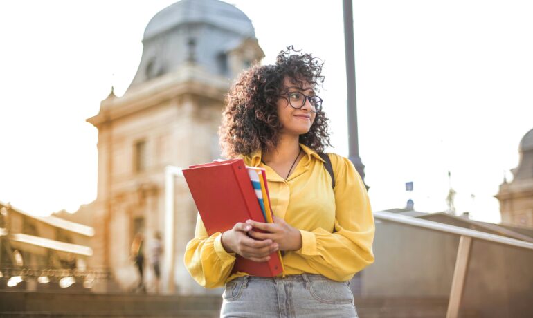 Smiling female student with curly hair and glasses holding books on campus steps.